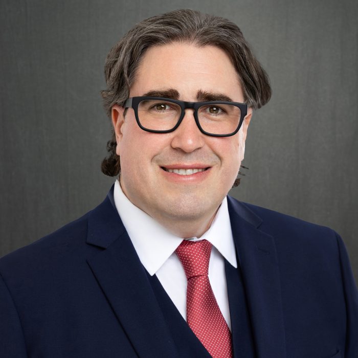 white male with glasses wearing a. navy blue suit and red tie