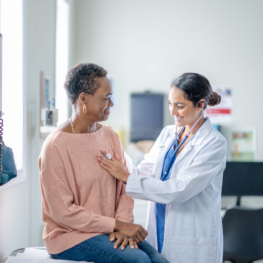 A senior woman of African decent sits up on an exam table during a routine check-up. She is sitting still and quiet as her female doctor listens to her heart.
